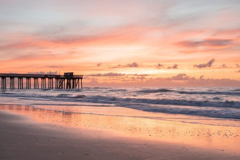 Sunrise over Avon Pier on Hatteras Island, with fishing rods lining the wooden pier and waves rolling onto the beach.