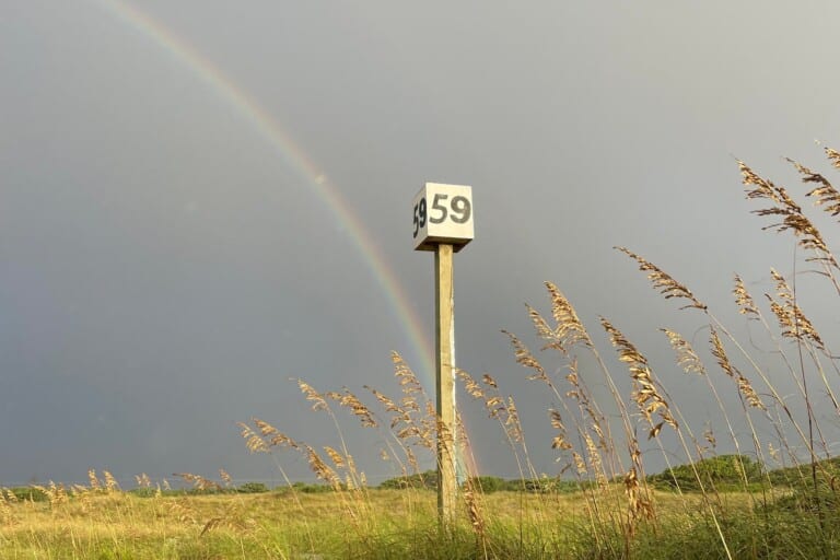 The numbered Ramp 59 marker post standing in sea oats with a rainbow over the dunes at Cape Hatteras National Seashore
