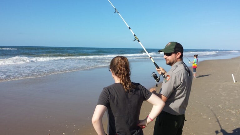 A park ranger teaching a beginner how to cast a surf rod at the water’s edge