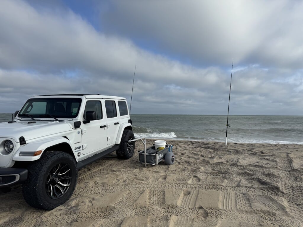 A white Jeep Wrangler parked beside surf fishing rods on the beach at the Outer Banks