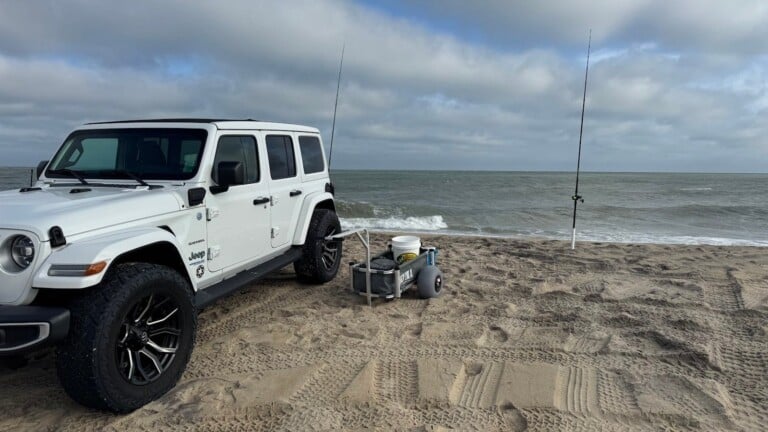 A white Jeep Wrangler parked on the sand beside a fishing cart and two surf rods in sand spikes at the water's edge — Outer Banks, NC