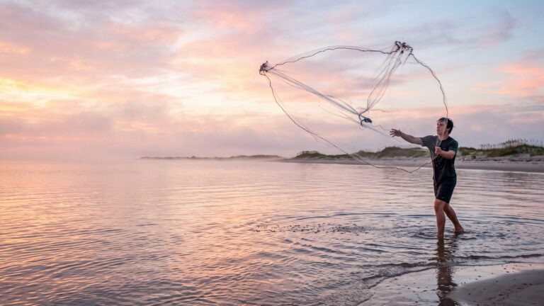 A young man throwing a cast net in a perfect circle over shallow sound water at sunrise — Outer Banks, NC