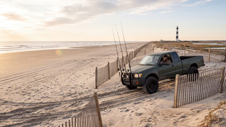 A 4x4 truck with surf rods on a front rack coming down a sandy beach ramp between dune fences, a banded lighthouse behind — Outer Banks, NC
