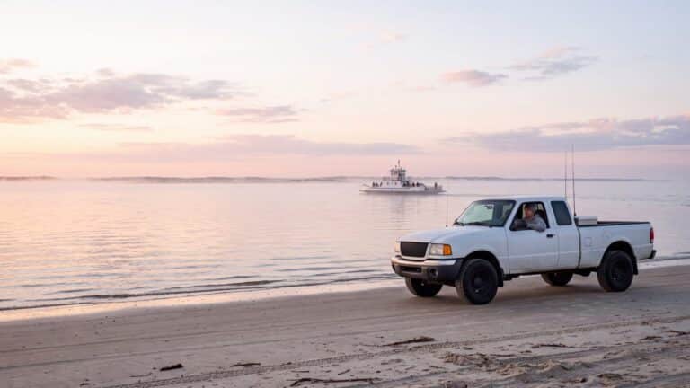 A 4x4 truck on a wide flat beach near an inlet, a small white ferry crossing far behind — Outer Banks, NC