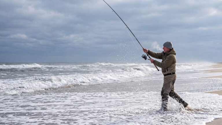 An angler in a knit cap and waders casting a long surf rod into cold grey surf, spray frozen in the air — Outer Banks, NC