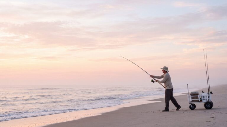 An angler in a wide-brim hat and sun shirt casting at first light before the summer crowd, a beach cart behind him — Outer Banks, NC