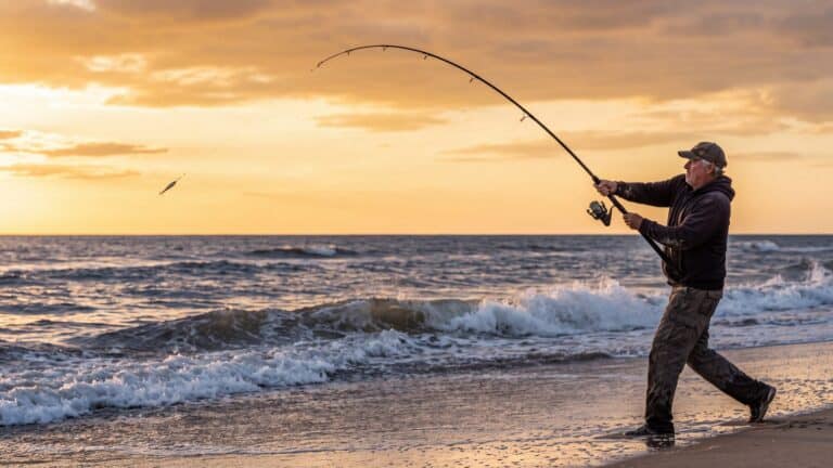 An angler loading a long surf rod into a deep bend on a cast, the rod tip whipping — Outer Banks, NC